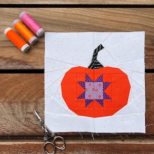 Puede incluir: Un bloque de calabaza de tela hecho a mano con un cuerpo naranja y una estrella morada en el centro. El tallo es negro y blanco. Tres carretes de hilo en naranja y rosa están a la izquierda. Un par de tijeras está en la parte inferior.