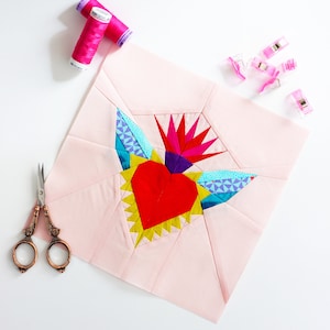 May include: A pink fabric square with a colorful heart design. The heart is made of fabric pieces in shades of red, yellow, blue, green, and purple. The heart is surrounded by a starburst pattern.