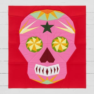 Könnte beinhalten: Ein pinkfarbener Sugar-Skull-Quiltblock auf rotem Hintergrund. Der Schädel besteht aus geometrischen Formen in Pink, Grün, Gelb und Orange. Ein dunkelgrüner Stern befindet sich auf der Stirn, und die Augen haben kreisförmige gelb-grüne Designs.