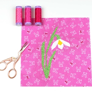May include: A pink fabric square with a white bunny rabbit pattern. A white and yellow paper flower with green stems is appliqued on the fabric. Three spools of pink thread are in the top left corner of the image.