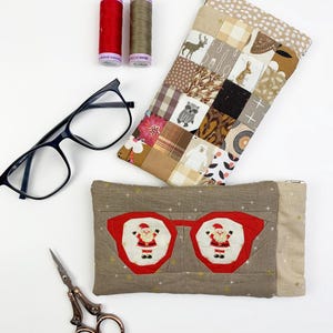 Può includere: Un assortimento di forniture e accessori per l'artigianato, tra cui un paio di occhiali neri, due rocchetti di filo e un paio di forbici decorative. Un astuccio per occhiali patchwork presenta un design di occhiali di Babbo Natale.