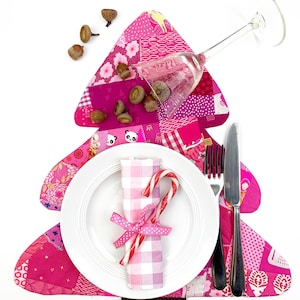 Puede incluir: Una mesa festiva con un mantel individual rosa en forma de árbol de retazos. Un plato blanco contiene una servilleta atada con una cinta rosa y bastones de caramelo. Una copa de vino y cubiertos están colocados en el mantel, con bellotas esparcidas.