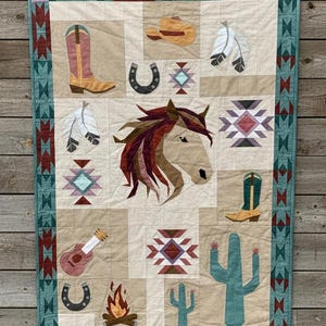 Op de afbeelding: Een patchworkquilt met een Western-thema. Het heeft een paardenkop, cowboylaarzen, een gitaar, een hoefijzer, een cactus en een kampvuur. De quilt heeft een turquoise rand en een neutrale achtergrond.