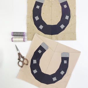 May include: Two fabric blocks with a horseshoe design in navy blue and gray, set against a tan background. The blocks are surrounded by sewing supplies, including thread spools in gray and white, and small scissors.