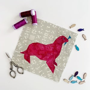 May include: A pink fabric seal with a fish in its mouth, surrounded by wooden fish cutouts. The seal is on a gray fabric background with a geometric pattern. There are three spools of purple thread in the upper left corner of the image.