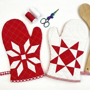Könnte beinhalten: Zwei gesteppte Ofenhandschuhe mit roten und weißen Schneeflocken- und Sterndesigns. Ein Paar Holzlöffel, eine Schere und Garnrollen sind ebenfalls vorhanden. Die Handschuhe haben eine rot-weiß karierte Bordüre.