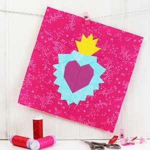 May include: A pink fabric square with a blue and white heart design and a yellow crown. The heart is pink and has a white outline. The crown is yellow and has a white outline. The fabric square is hanging from a white string with two yellow clothespins.