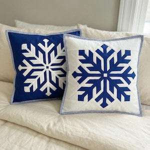 Op de afbeelding: Twee vierkante kussens met een sneeuwvlokontwerp. Eén kussen heeft een witte sneeuwvlok op een marineblauwe achtergrond, de andere een marineblauwe sneeuwvlok op een witte achtergrond. Beide kussens hebben een gestreepte rand.