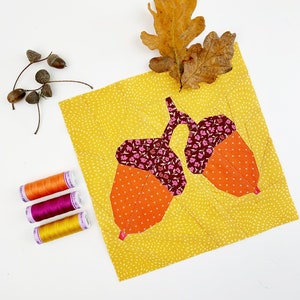 May include: A yellow fabric square with a brown, orange and pink acorn design. Three spools of thread are on the right side of the image. The thread colours are orange, pink and yellow.