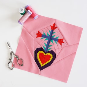 May include: A pink fabric square with a colorful heart and flower design. The fabric is folded in half and there are three spools of thread and a pair of scissors on the white surface.