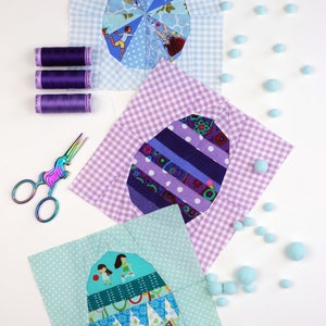 May include: Three fabric patchwork squares with different designs of colorful eggs. Each square is surrounded by a white gingham border. The squares are arranged in a triangle shape on a white surface. There are also small blue pom-poms scattered around the squares.