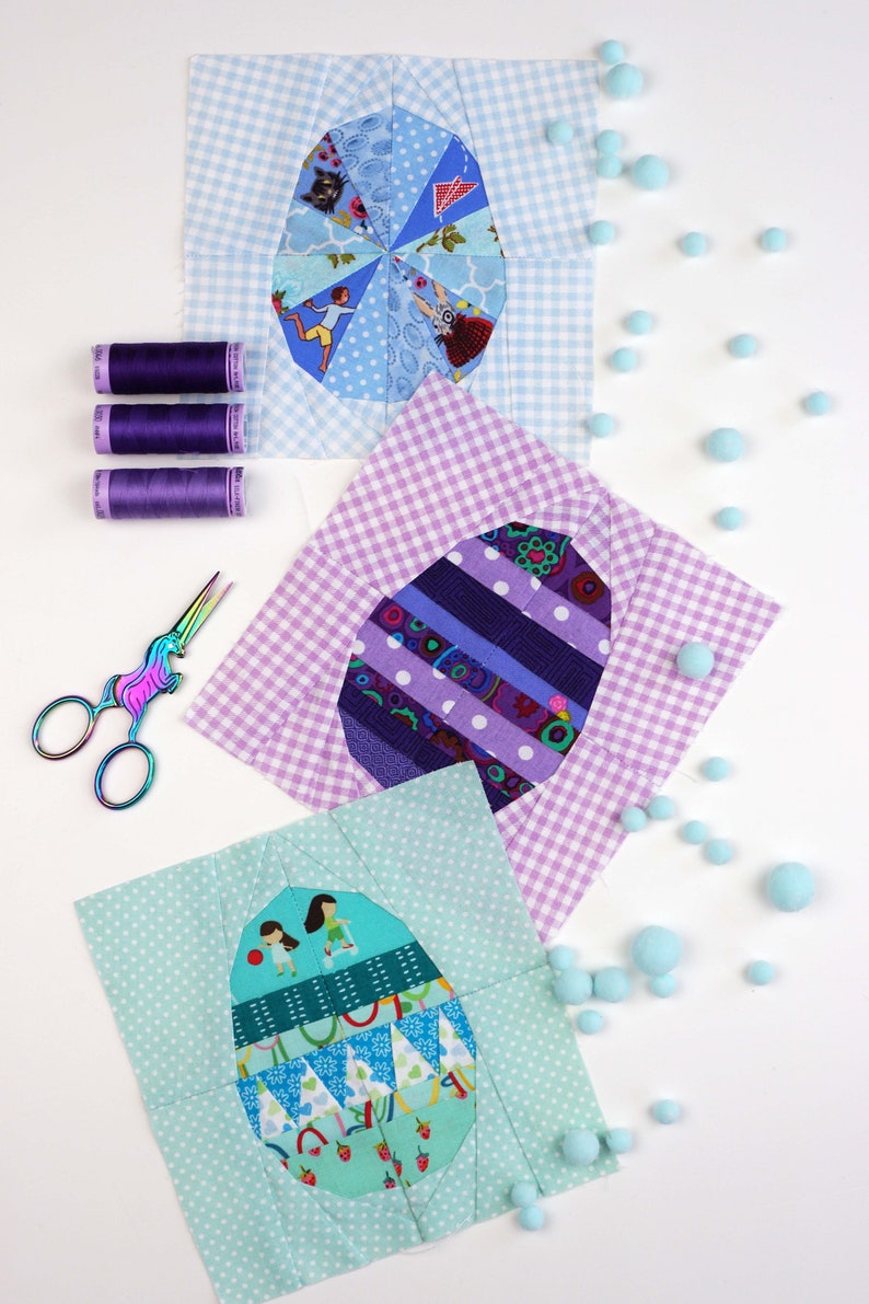 May include: Three fabric patchwork squares with different designs of colorful eggs. Each square is surrounded by a white gingham border. The squares are arranged in a triangle shape on a white surface. There are also small blue pom-poms scattered around the squares.