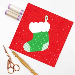 Peut inclure: Un fond en feutre rouge avec une applique de chaussette de Noël en feutre blanc et vert. La chaussette a un revers en feutre blanc et un corps en feutre vert avec des pois blancs. Il y a deux bobines de fil blanc et une paire de ciseaux dorés sur une surface blanche.