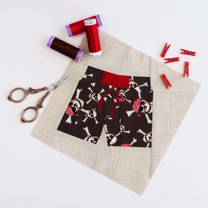 May include: A fabric pirate-themed shorts applique with a brown skull and crossbones pattern, red waistband, and red drawstrings. The applique is on a beige background, with red thread spools, red clothespins, and vintage scissors.