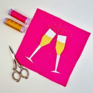 May include: Two white champagne glasses with yellow liquid and gold confetti on a bright pink background. Two spools of thread are visible in the upper left corner of the image.