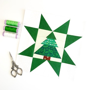 May include: A festive Christmas tree quilt block featuring a green star design and a patterned tree. Three spools of green and white thread and a pair of scissors are placed beside the quilt block, ready for sewing.