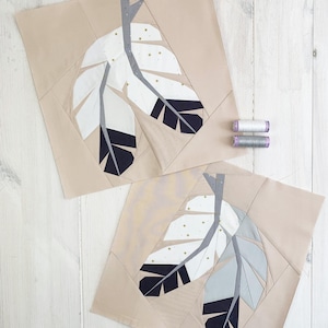 May include: Two beige fabric squares featuring feather designs. The feathers are white, grey, and navy blue, with gold star accents. Two spools of thread are next to the squares. The background is a white wooden surface.