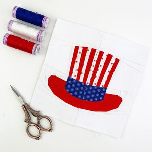 Può includere: Un quadrato di tessuto bianco con un cappello di tessuto di zio Sam rosso, bianco e blu cucito sopra. Tre bobine di filo sono nell'angolo in alto a sinistra dell'immagine: rosso, bianco e blu.
