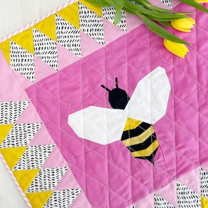 May include: A pink and yellow quilt with a black and yellow bee design in the center. The quilt is made of various geometric shapes and patterns.
