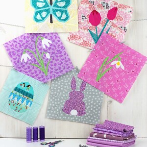 May include: A set of six colorful fabric squares with various spring-themed designs, including a butterfly, flowers, and an egg. The squares are hanging on a white wooden background with clothespins. There are also spools of purple thread and a pair of scissors on a white surface.