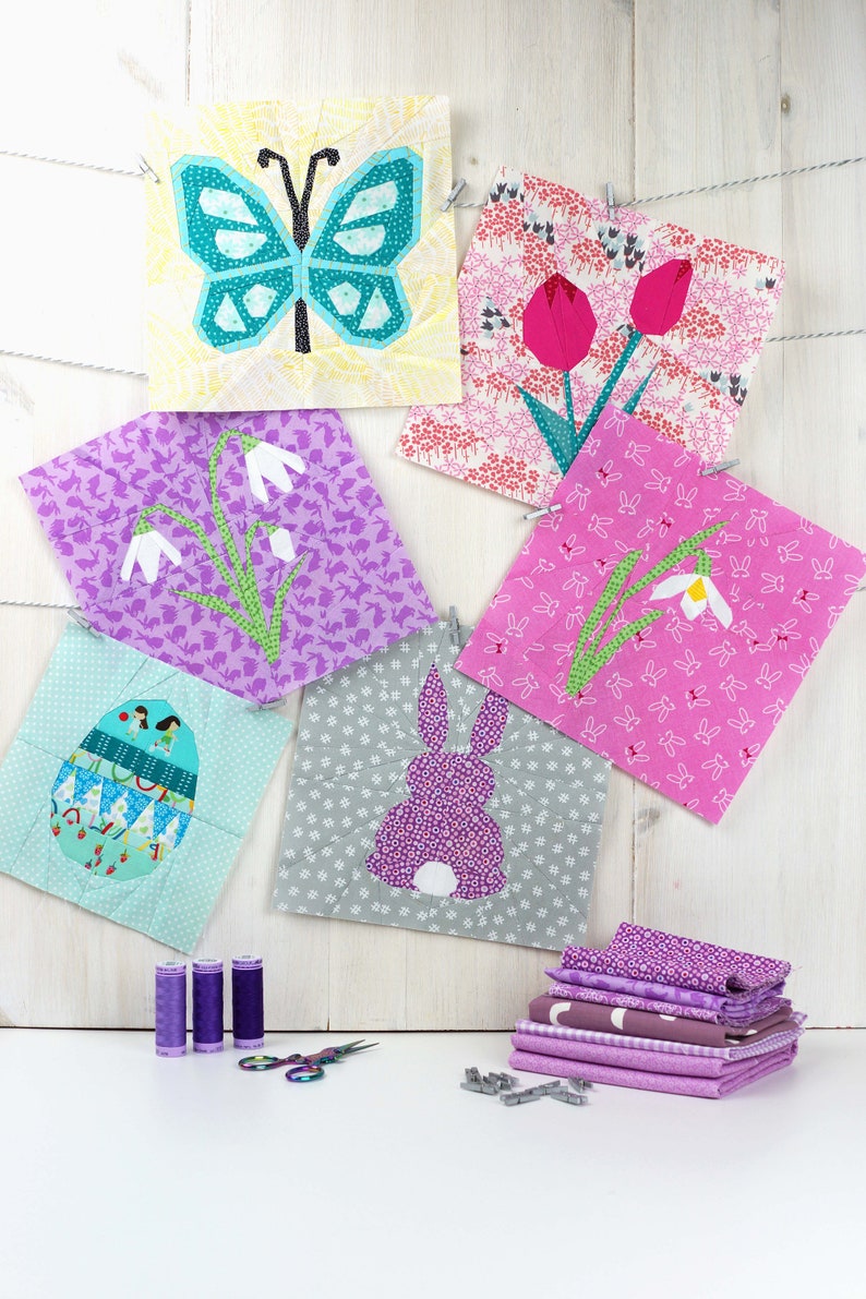 May include: A set of six colorful fabric squares with various spring-themed designs, including a butterfly, flowers, and an egg. The squares are hanging on a white wooden background with clothespins. There are also spools of purple thread and a pair of scissors on a white surface.