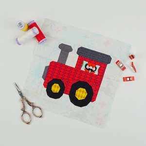 May include: A red and black fabric train applique with a yellow wheel and a small figure of a person with a mustache in the driver's seat. The applique is on a white background with a small, colorful pattern.