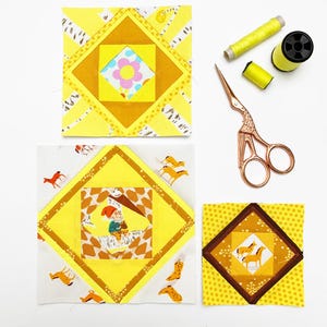 May include: Three yellow and patterned quilt blocks with a flower, a gnome, and horses. Also, a pair of rose gold scissors, and spools of yellow thread are visible. The quilt blocks are square and feature a diamond design.