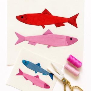 May include: Three pieces of art featuring origami fish in red, pink, and blue. The fish are on off-white backgrounds. Gold scissors and pink thread spools are also in the image. The fish art is a craft project.