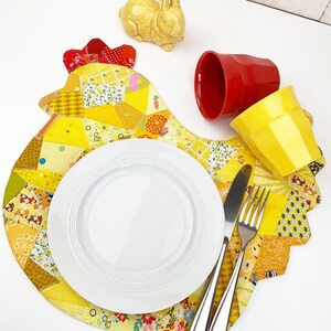 May include: A yellow and patchwork placemat shaped like a hen, with a white plate, silverware, and two cups. A yellow ceramic bunny figurine and a red cup are also on the white surface. The placemat has a red comb.