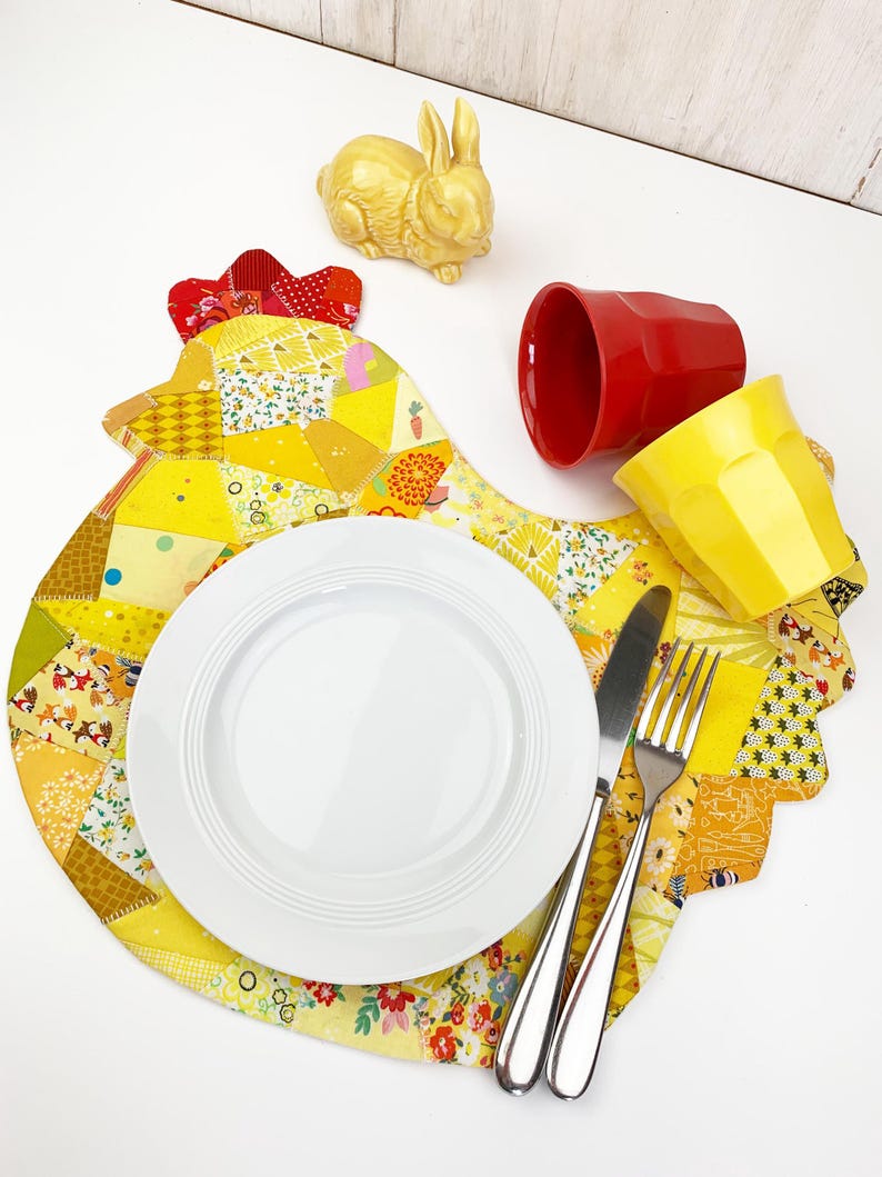 May include: A yellow and patchwork placemat shaped like a hen, with a white plate, silverware, and two cups. A yellow ceramic bunny figurine and a red cup are also on the white surface. The placemat has a red comb.
