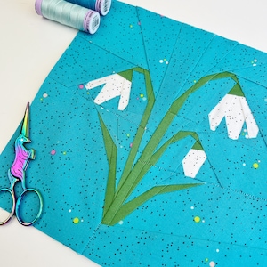 Könnte beinhalten: Ein blauer Stoffquadrat mit weißen Schneeglöckchen und grünen Stielen. Der Stoff hat ein weißes Punkte-Muster. Zwei Garnrollen befinden sich in der oberen linken Ecke des Bildes.