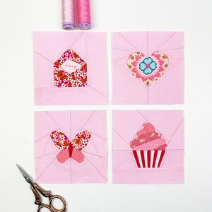May include: Four pink fabric squares with appliqued designs: an envelope with "I Love You" text, a heart, a butterfly, and a cupcake. Two spools of pink thread and a pair of scissors are also present. The items are arranged on a white surface.