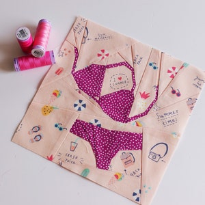 May include: A pink and peach fabric patchwork quilt block with a summer theme. The block features a pink swimsuit, ice cream cones, pineapples, sunglasses, flip flops, a camera, a backpack, and the text "Summer time!" and "Fresh Juice".
