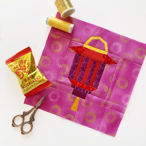 May include: A pink fabric square with a gold and red paper lantern design. The fabric has a white polka dot pattern. There is a spool of gold thread, a spool of yellow thread, a pair of scissors, and a packet of fortune cookies on a white surface.