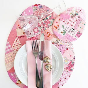 May include: A charming table setting with a pink patchwork placemat, matching coaster, and a white plate. Silverware and a pink napkin are arranged on the plate, with two bunny-shaped napkin rings. A clear wine glass rests on the placemat.
