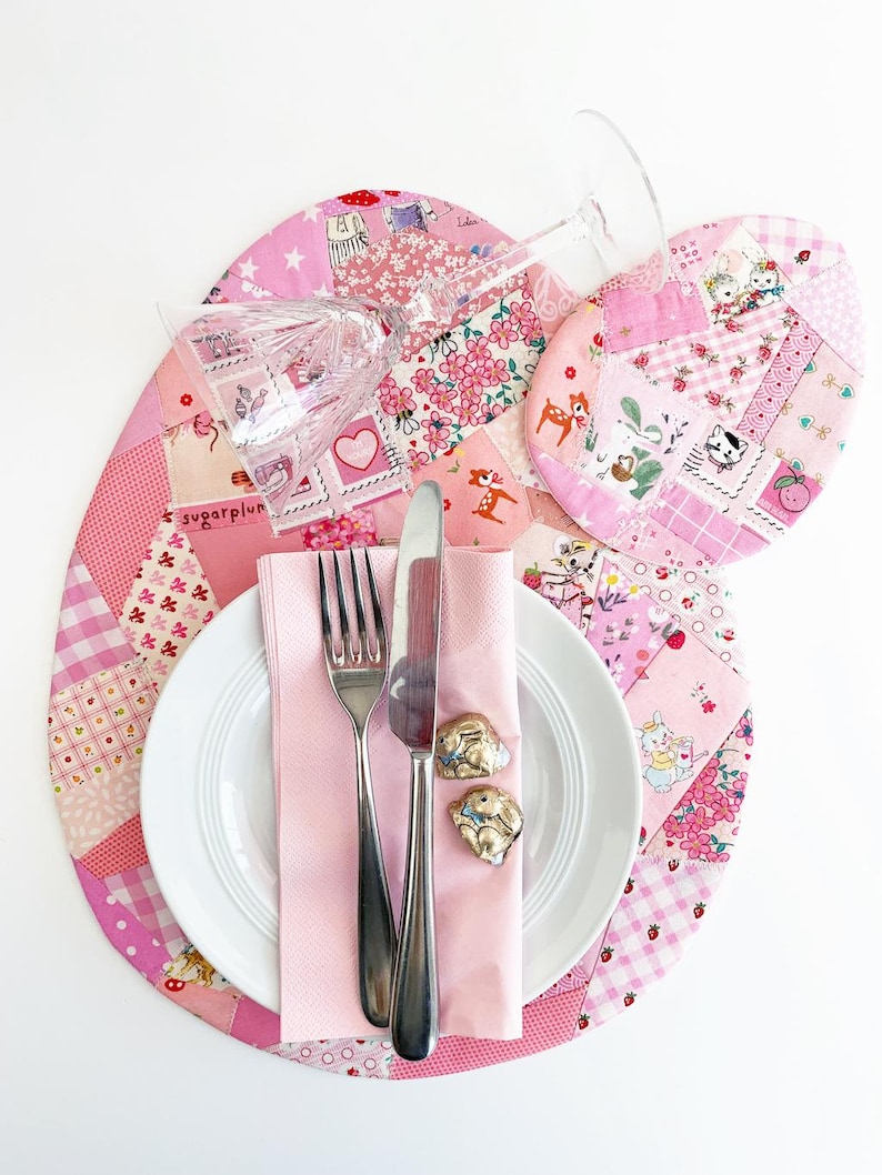 May include: A charming table setting with a pink patchwork placemat, matching coaster, and a white plate. Silverware and a pink napkin are arranged on the plate, with two bunny-shaped napkin rings. A clear wine glass rests on the placemat.