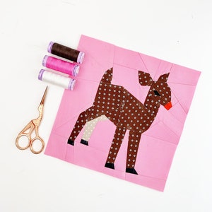 May include: A pink fabric square with a brown deer applique with white polka dots. The deer has a red nose and black hooves. There are three spools of thread in pink, white, and brown, and a pair of gold scissors on a white surface.