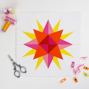 May include: A quilt block with a starburst design in shades of yellow, orange, pink, and red. The block is pinned to a white surface with quilting clips.