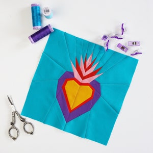May include: A turquoise fabric square with a colorful heart design in the center. The heart is made of purple, red, yellow, and pink fabric. There are sewing clips and spools of thread on the white surface.