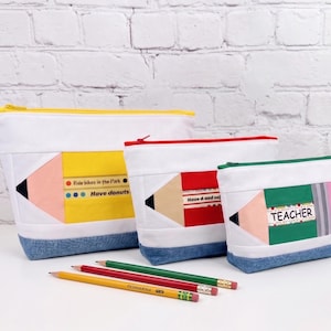 Puede incluir: Tres estuches con cremallera en forma de lápiz en amarillo, rojo y verde, cada uno con base de mezclilla azul y parte superior blanca. El estuche amarillo tiene el texto Ride bikes in the Park y Have donuts. El verde tiene la palabra TEACHER.