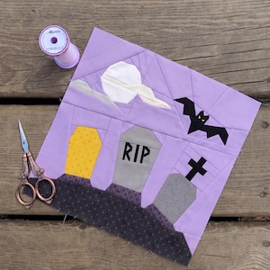 May include: A square fabric quilt block featuring a graveyard scene with a yellow coffin, two grey tombstones, one marked with RIP, a black cross, a white moon, and a black bat against a light purple sky, accompanied by scissors and thread.