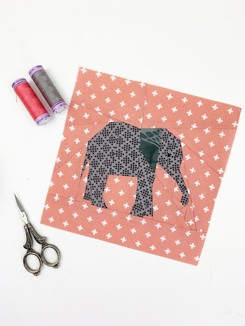 May include: A gray and green elephant fabric applique on a pink fabric background with a white plus sign pattern. The fabric is cut out and ready to be sewn.