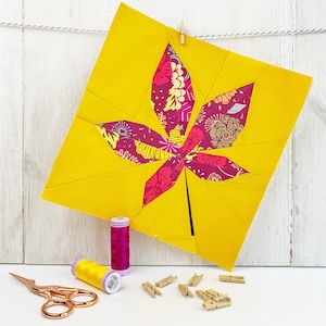 Op de afbeelding: Een geel stoffen blad met een roze en wit bloemenpatroon erin. Het blad hangt aan een touwtje met een houten wasknijper. Er zijn twee spoelen garen, een gele en een roze, en een aantal houten wasknijpers op een wit oppervlak.