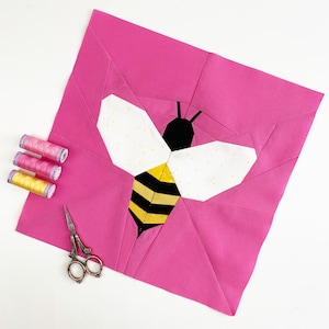 Könnte beinhalten: Ein rosa Stoffquadrat mit einem schwarzen und gelben Bienendesign in der Mitte. Die Biene hat weiße Flügel mit gelben Punkten. Der Stoff ist zu Dreiecken gefaltet, wodurch ein geometrisches Muster entsteht.