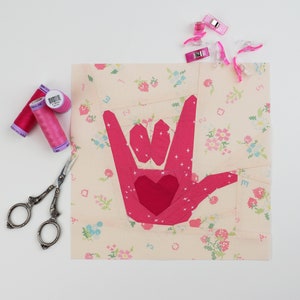 Peut inclure: Une applique en tissu rose du symbole de la langue des signes américaine pour "Je t'aime" sur un fond blanc avec un motif floral rose. L'applique est faite de plusieurs morceaux de tissu cousus ensemble.