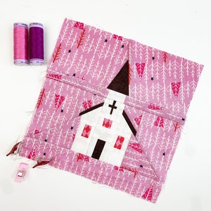 May include: A pink fabric block with a white and brown church applique. The fabric has a white and pink tree pattern.