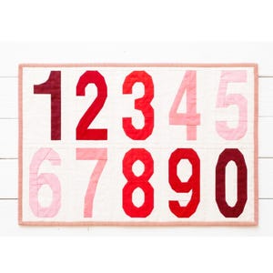 May include: A quilted number chart featuring the numbers 0 through 9 in various shades of red, from deep burgundy to light pink. The numbers are set against a cream-colored background and framed with a red and white striped border. The quilt is rectangular.