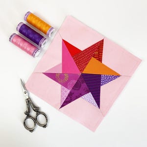 May include: A pink fabric star with a multi-colored patchwork design. The star is made up of various shades of pink, purple, orange, and red fabric. There are three spools of thread in the image, one orange, one purple, and one pink.