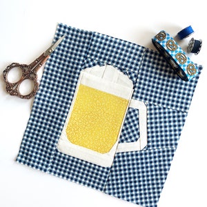 Puede incluir: Un cuadrado de tela de cuadros azules y blancos con una aplicación blanca y amarilla de una jarra de cerveza. La tela está doblada por la mitad y hay artículos de costura en la mesa.