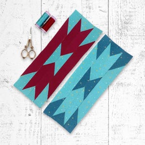May include: Two fabric strips with a geometric design in teal and burgundy, with gold scissors and spools of thread. The strips have a chevron pattern. The background is a white wooden surface. Sewing and quilting supplies.